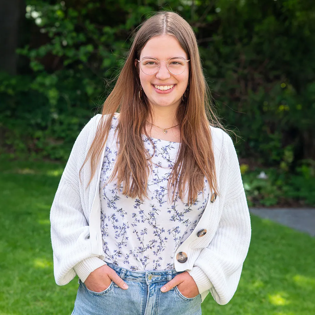 Das Bild zeigt eine strahlende junge Frau in selbstbewusster Pose vor einem natürlichen grünen Hintergrund in Schleswig – als professionelles und ausdrucksstarkes Bewerbungsfoto.