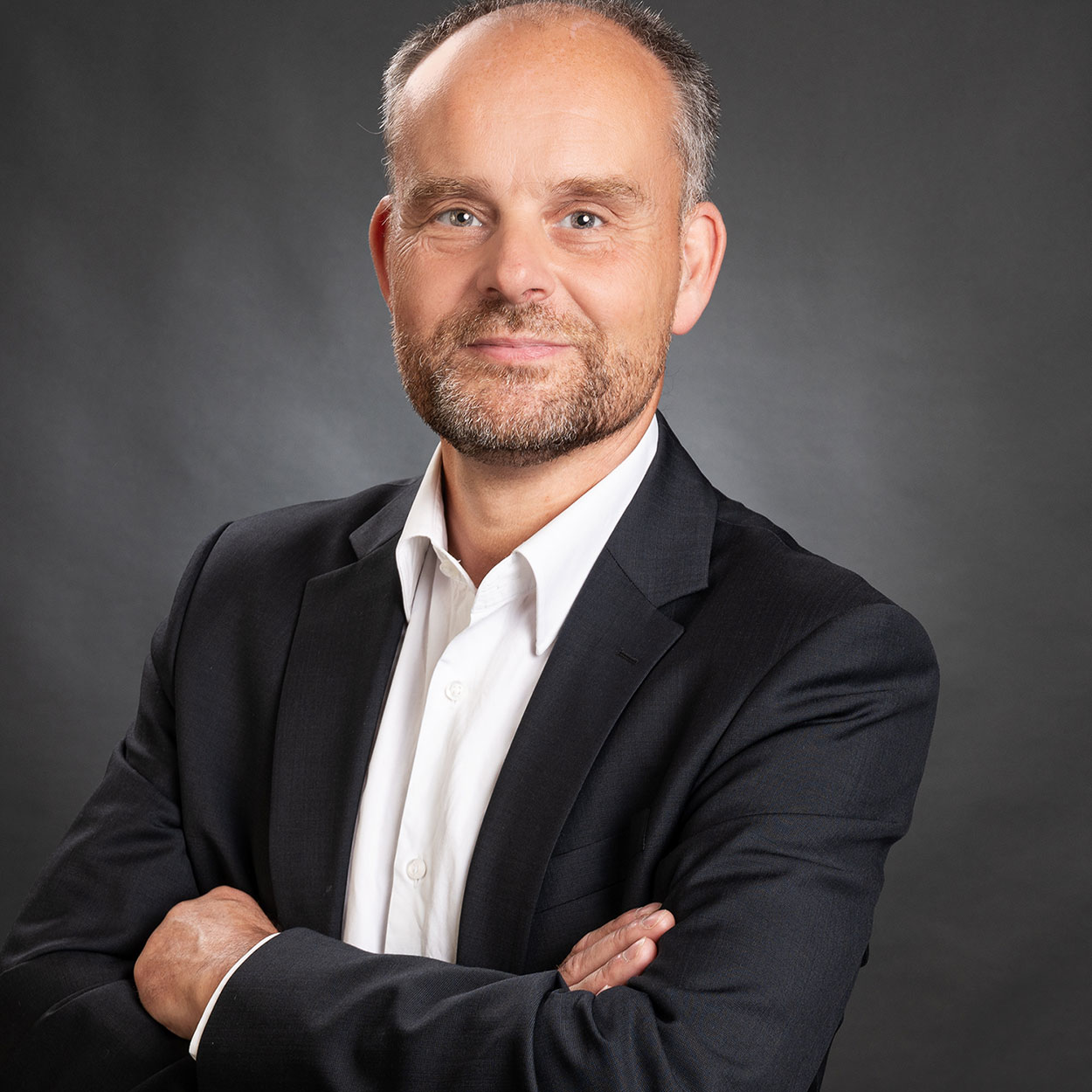 Business-Porträt eines älteren Mannes mit verschränkten Armen, weißem Hemd und schwarzem Sakko vor anthrazitfarbenem Hintergrund in Schleswig, mit ernstem Blick und leichtem Schmunzeln.